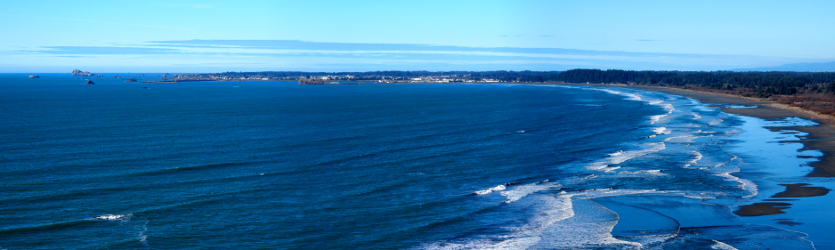 [Several photos stitched together showing the extension of the land into the ocean in the far distance and the land hooking back around to the right to near where the photo was taken. The image is mostly water as the waves come to shore at low tide.]